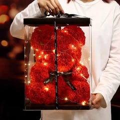 Red rose teddy bear with lights in a clear box held by a person.