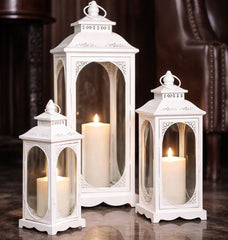 Set of three white lanterns with candles inside on a reflective surface.