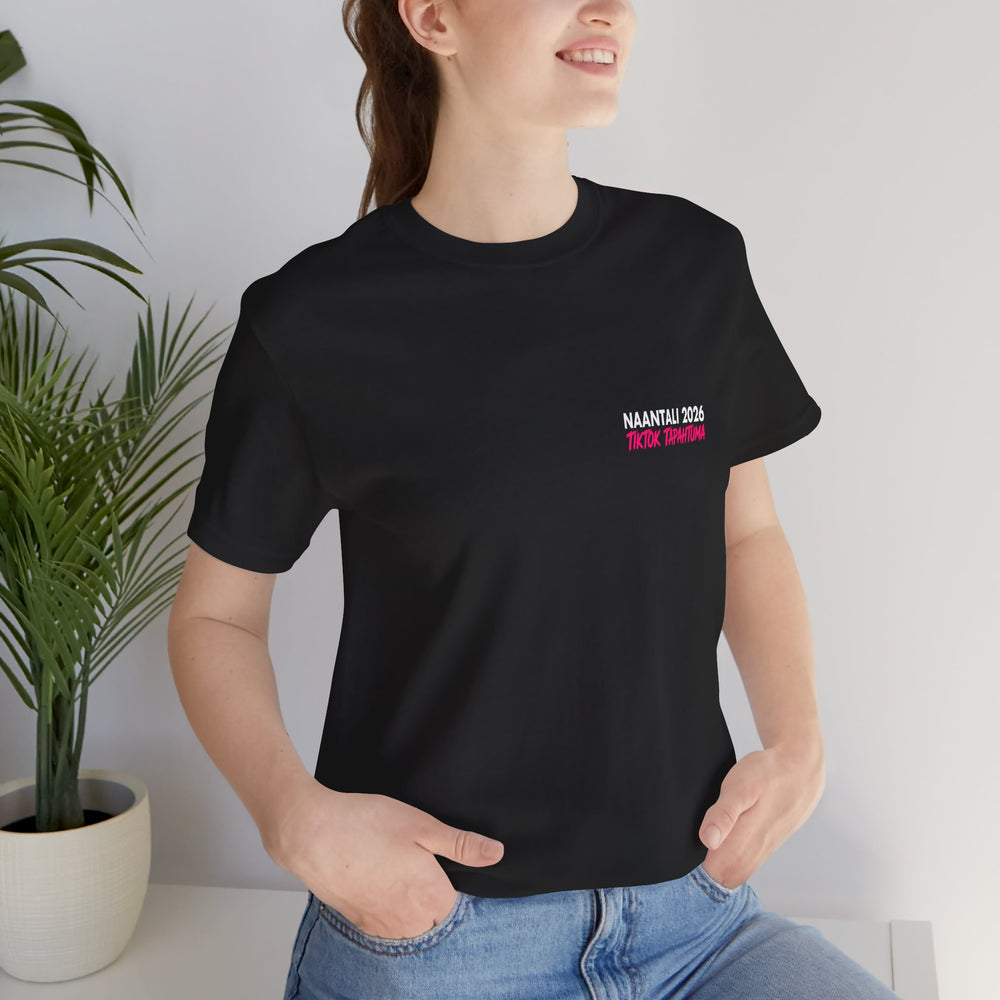 Person wearing a black t-shirt with a text, standing indoors with a plant in the background.