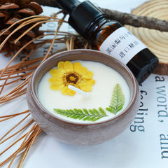 Candle with a yellow flower and green leaf in a brown pot, next to a bottle of essential oil on a newspaper background.