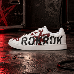 A white low-top sneaker with bold black 'ROKROK' lettering and red graphic accents on the side, photographed on a dark, wet stage floor with equipment and red lighting in the background.