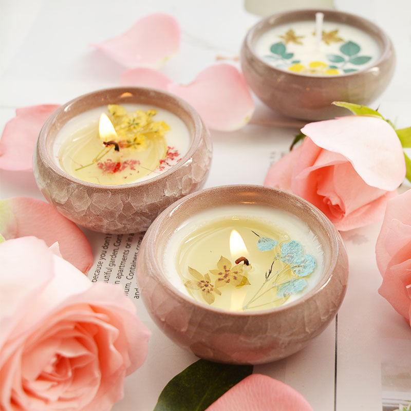 Three small candles with floral designs in ceramic holders surrounded by pink roses on a light wooden surface.