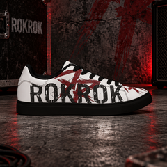 A white low-top sneaker with black and red graphic details, featuring the word 'ROKROK' printed across the side, set against a dark, industrial stage background with red lighting and audio equipment.