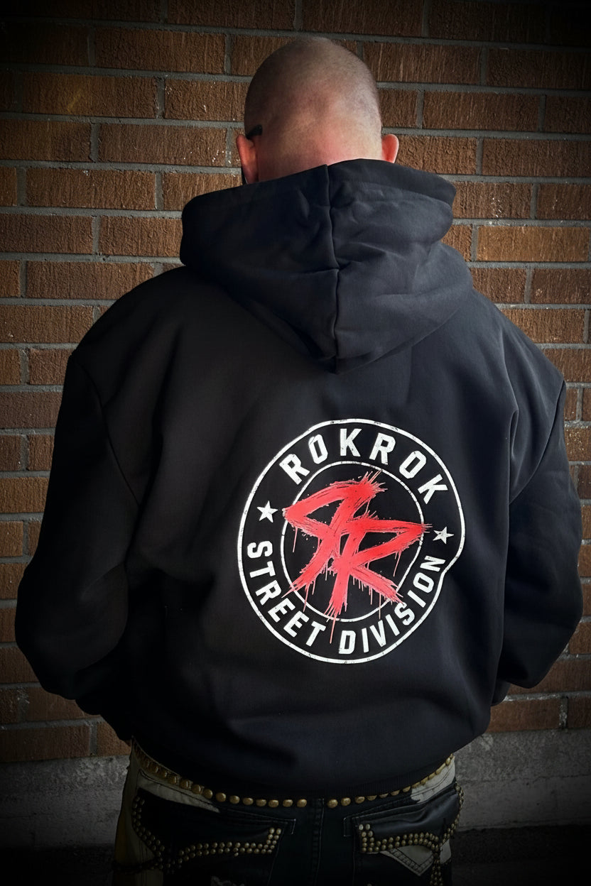 A man viewed from behind wearing a black pullover hoodie with a large circular back graphic that reads 'ROKROK STREET DIVISION' around a red stylized 'RR' logo, standing against a brick wall.