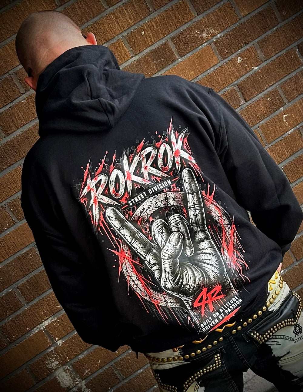 A man viewed from behind wearing a black hoodie with a large red, white, and gray graphic on the back that reads 'ROKROK' and 'STING CHAMPIONSHIP'. He is standing against a brown brick wall.