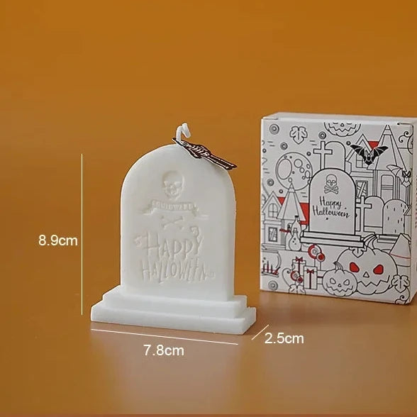 A Halloween tombstone candle shown next to its illustrated box on an orange-brown background. The candle is white with an embossed 'Happy Halloween' design and a small skull-and-bow detail on top.