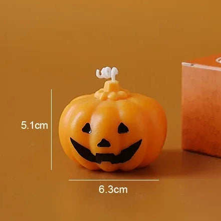 An orange jack-o'-lantern-shaped candle shown next to its brown cardboard box on a matching orange-brown background, with dimension labels indicating 5.1 cm height and 6.