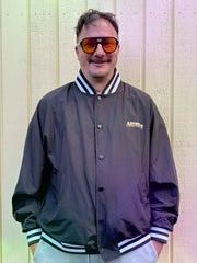 A man wearing a black bomber jacket with white striped ribbed collar, cuffs, and hem, along with orange-tinted sunglasses, standing against a light-colored wooden wall.