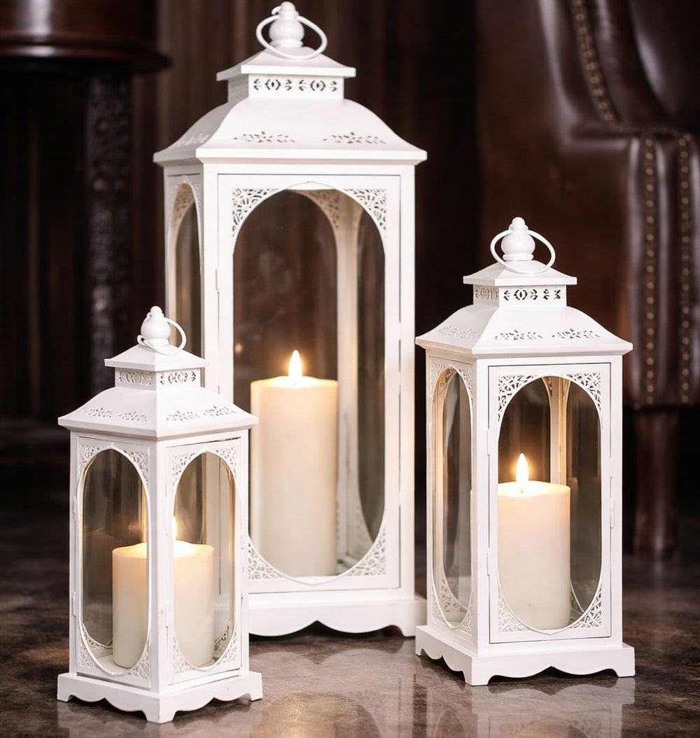 Set of three white lanterns with candles inside on a reflective surface.