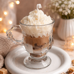 Hot chocolate with whipped cream in a clear glass mug on a white plate, surrounded by cinnamon sticks and sugar cubes.