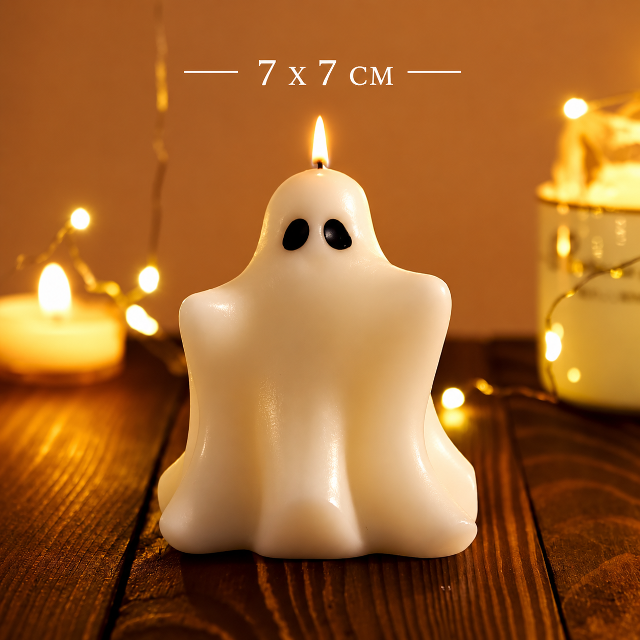 A ghost-shaped white candle with black eyes, lit and placed on a wooden surface with warm string lights in the background. White text at the top reads '7 x 7 CM'.